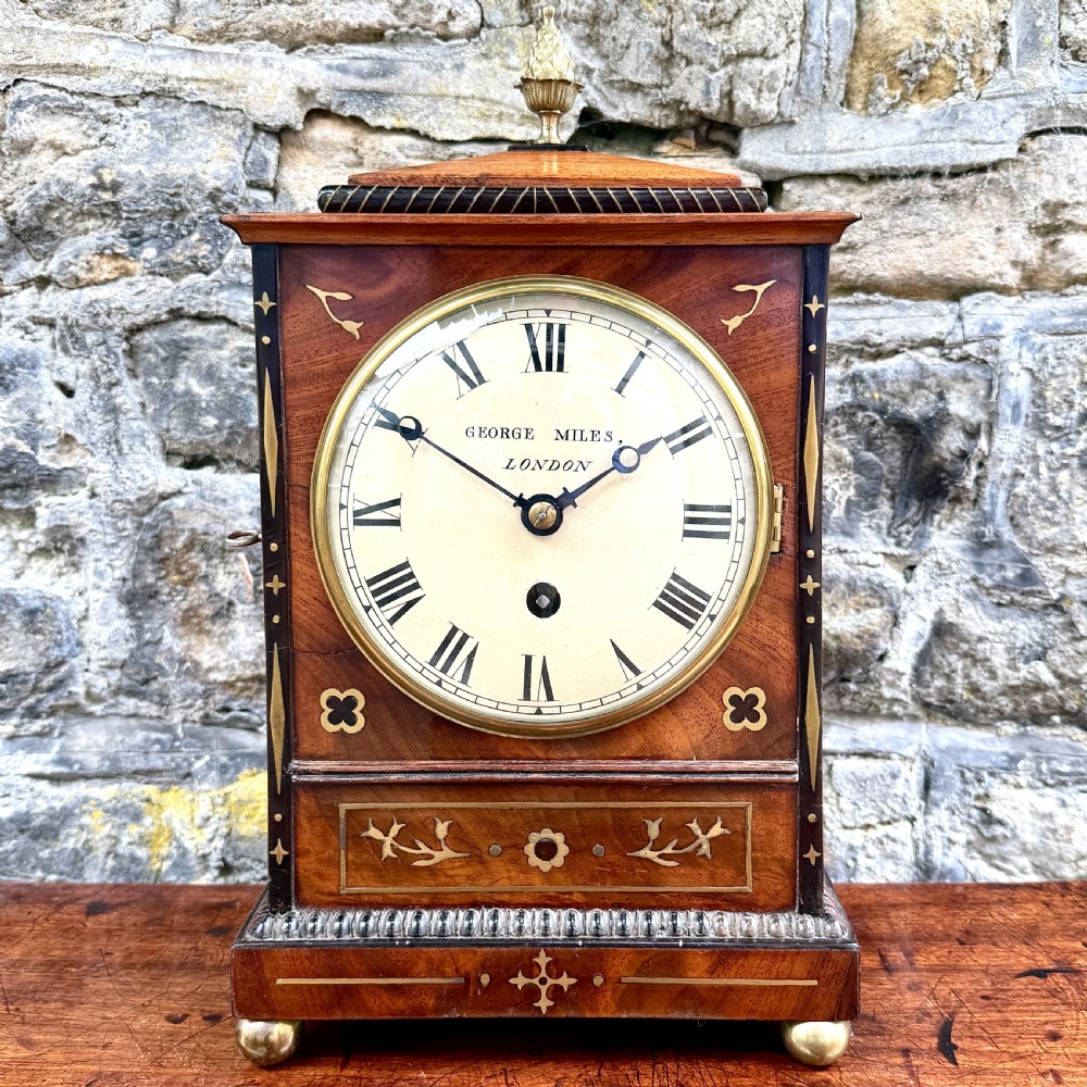 A Regency mahogany library clock featuring brass floral inlay, a pineapple finial, brass ball feet, and a white dial signed George Miles London.