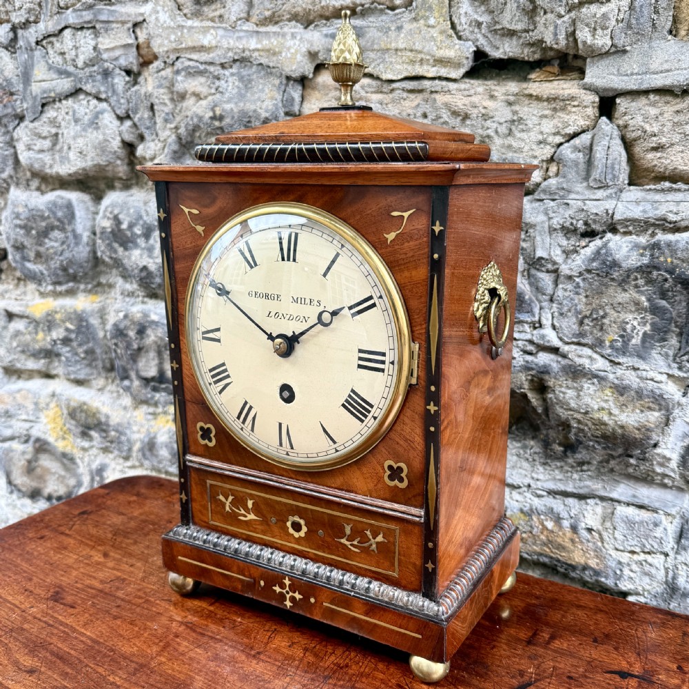 A Regency mahogany library clock featuring brass floral inlay, a pineapple finial, brass ball feet, and a white dial signed George Miles London. View II