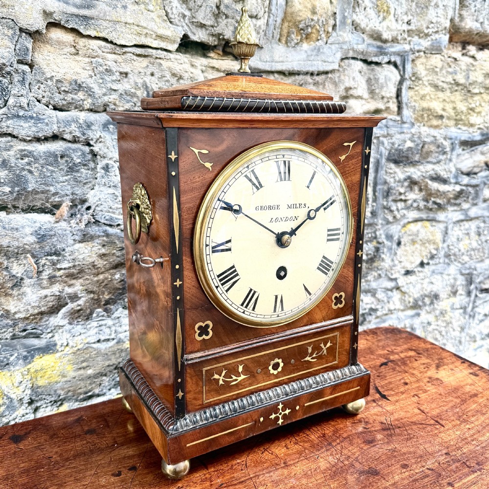 A Regency mahogany library clock featuring brass floral inlay, a pineapple finial, brass ball feet, and a white dial signed George Miles London. View III