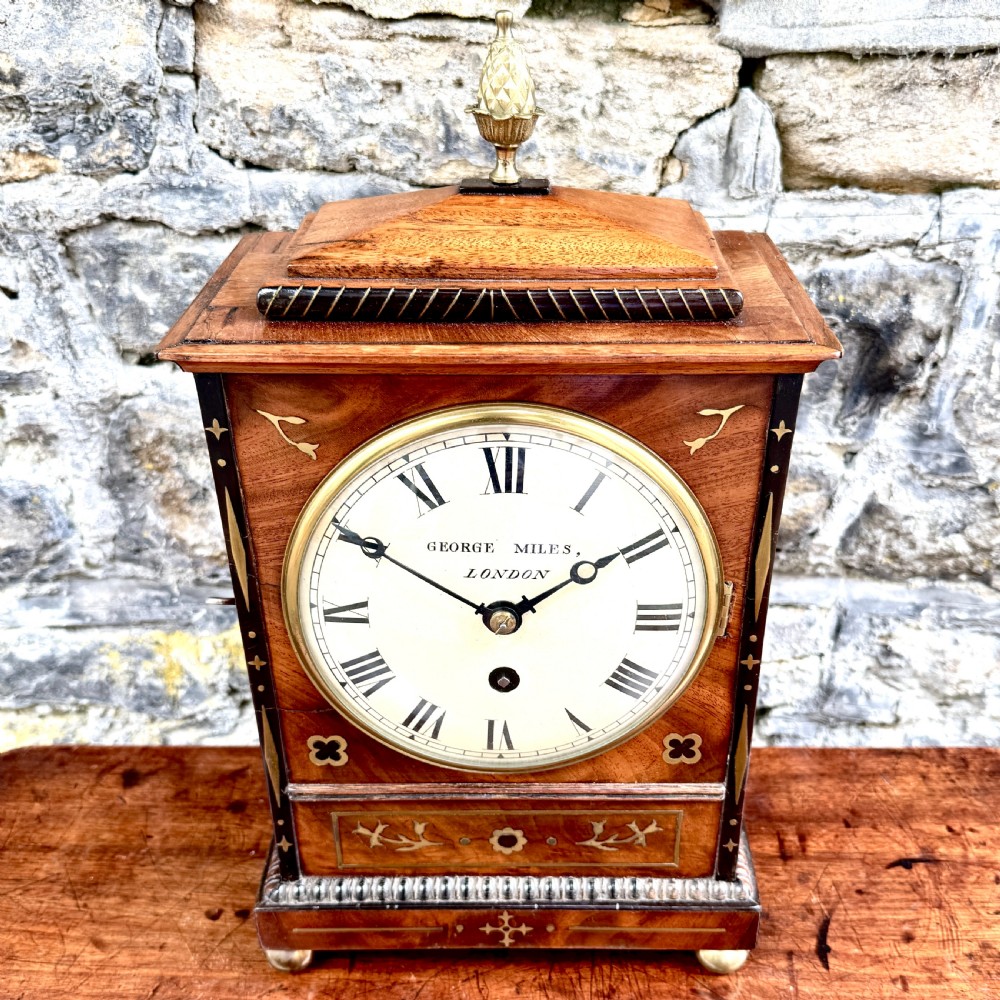A Regency mahogany library clock featuring brass floral inlay, a pineapple finial, brass ball feet, and a white dial signed George Miles London. View V