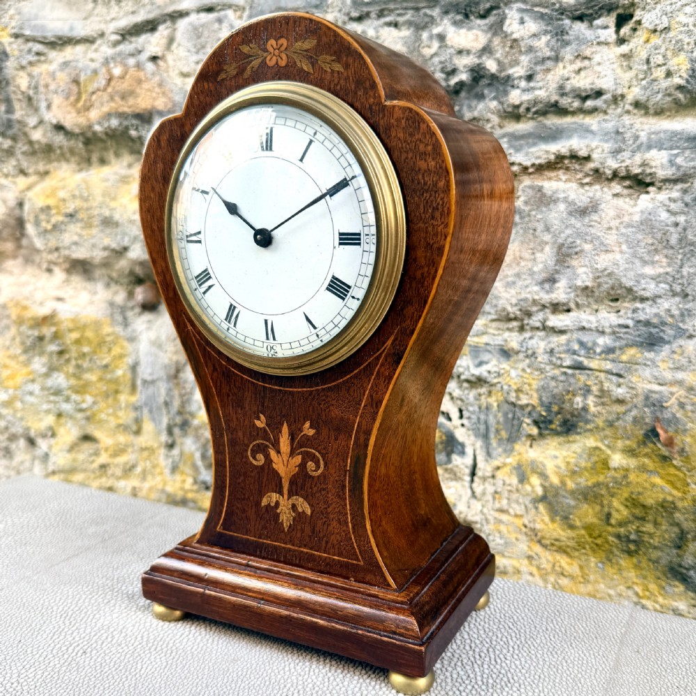 An Art Nouveau inlaid mahogany mantel clock featuring foliate marquetry, a white enamel dial with Roman numerals, and brass bun feet. View II