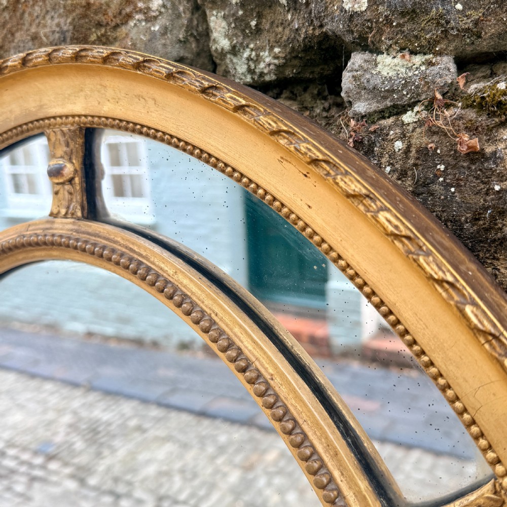 An oval giltwood wall mirror featuring a double-ring frame connected by small decorative spacers and decorated with beaded moulding. View II