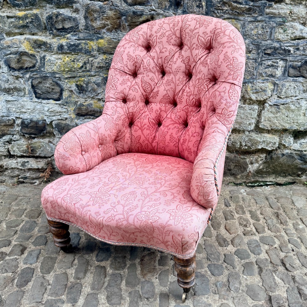 A mid-19th century French low bedroom chair featuring pink patterned button-tufted upholstery and turned wooden front legs on castors.
