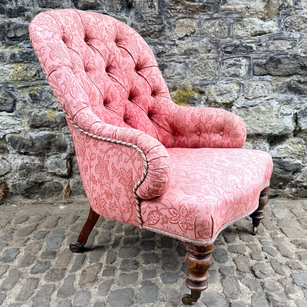 A mid-19th century French low bedroom chair featuring pink patterned button-tufted upholstery and turned wooden front legs on castors. View IV