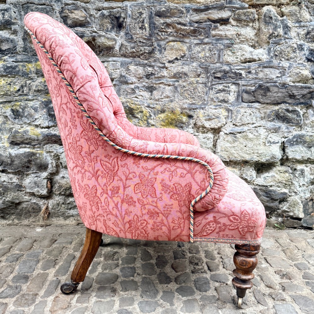 A mid-19th century French low bedroom chair featuring pink patterned button-tufted upholstery and turned wooden front legs on castors. View V