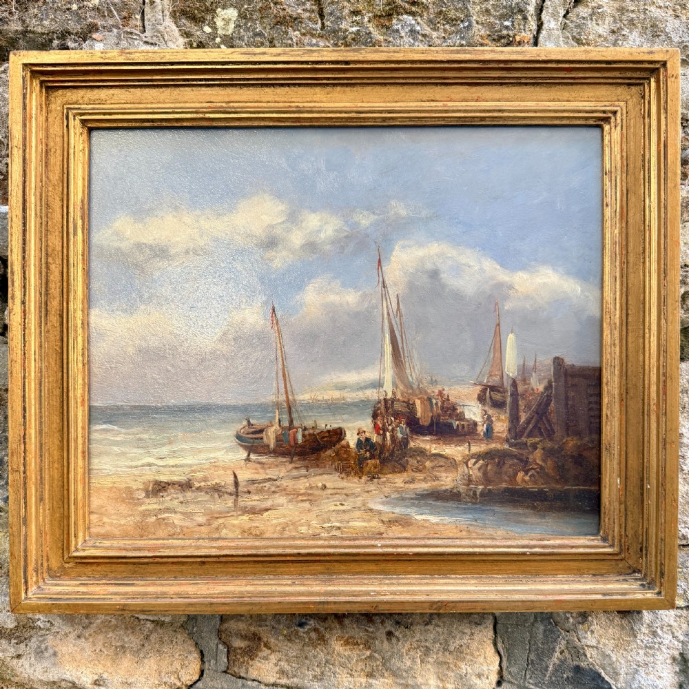 A nineteenth-century coastal oil painting depicting figures and fishing boats on a sandy shore under a cloudy sky, presented in a moulded gold frame.