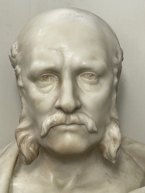 fig 5. A carved off-white marble bust depicting a bald male figure with a prominent moustache wearing classical draped fabric across the shoulders.