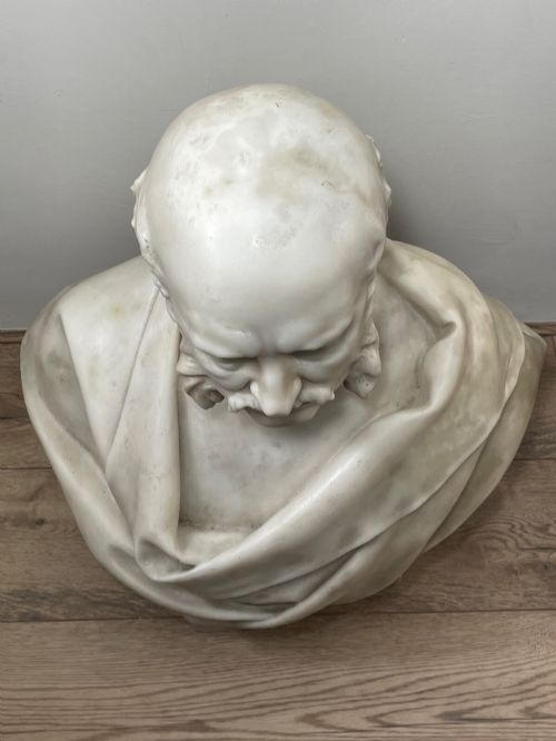 fig 8. A carved off-white marble bust depicting a bald male figure with a prominent moustache wearing classical draped fabric across the shoulders.