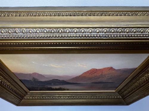 fig 9. An antique oil painting depicting a lake surrounded by mountains at sunset, enclosed within a carved gold frame.
