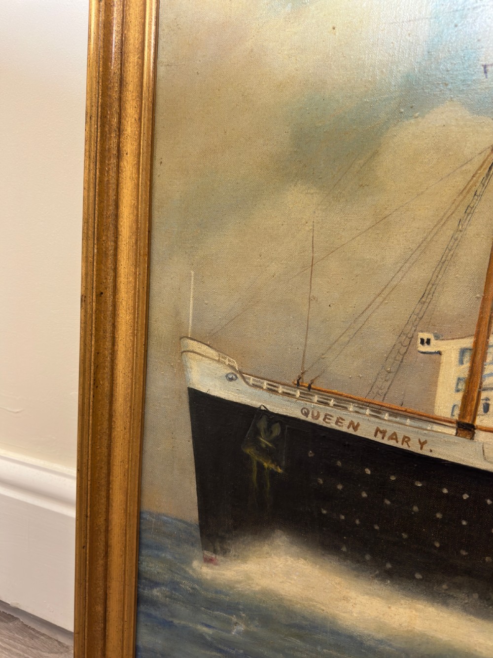 An oil painting depicting the RMS Queen Mary ocean liner sailing across blue waters under a cloudy sky, framed in a moulded gold gilt border. View V