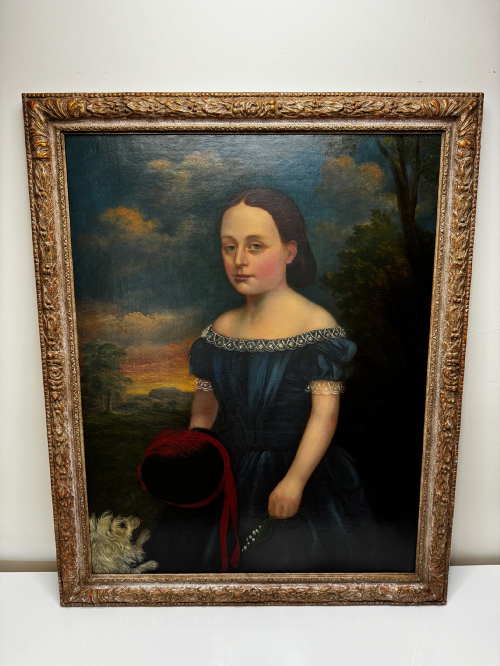 A Victorian oil portrait depicting a young girl in a dark blue off-the-shoulder dress holding a bonnet, accompanied by a small white dog, framed in an ornate gilded border. View II