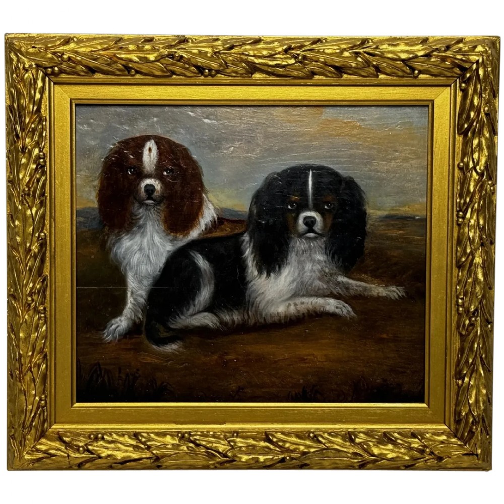A nineteenth-century oil painting depicting two King Charles Spaniels resting outdoors, enclosed within an ornate gilded frame decorated with carved laurel leaves.