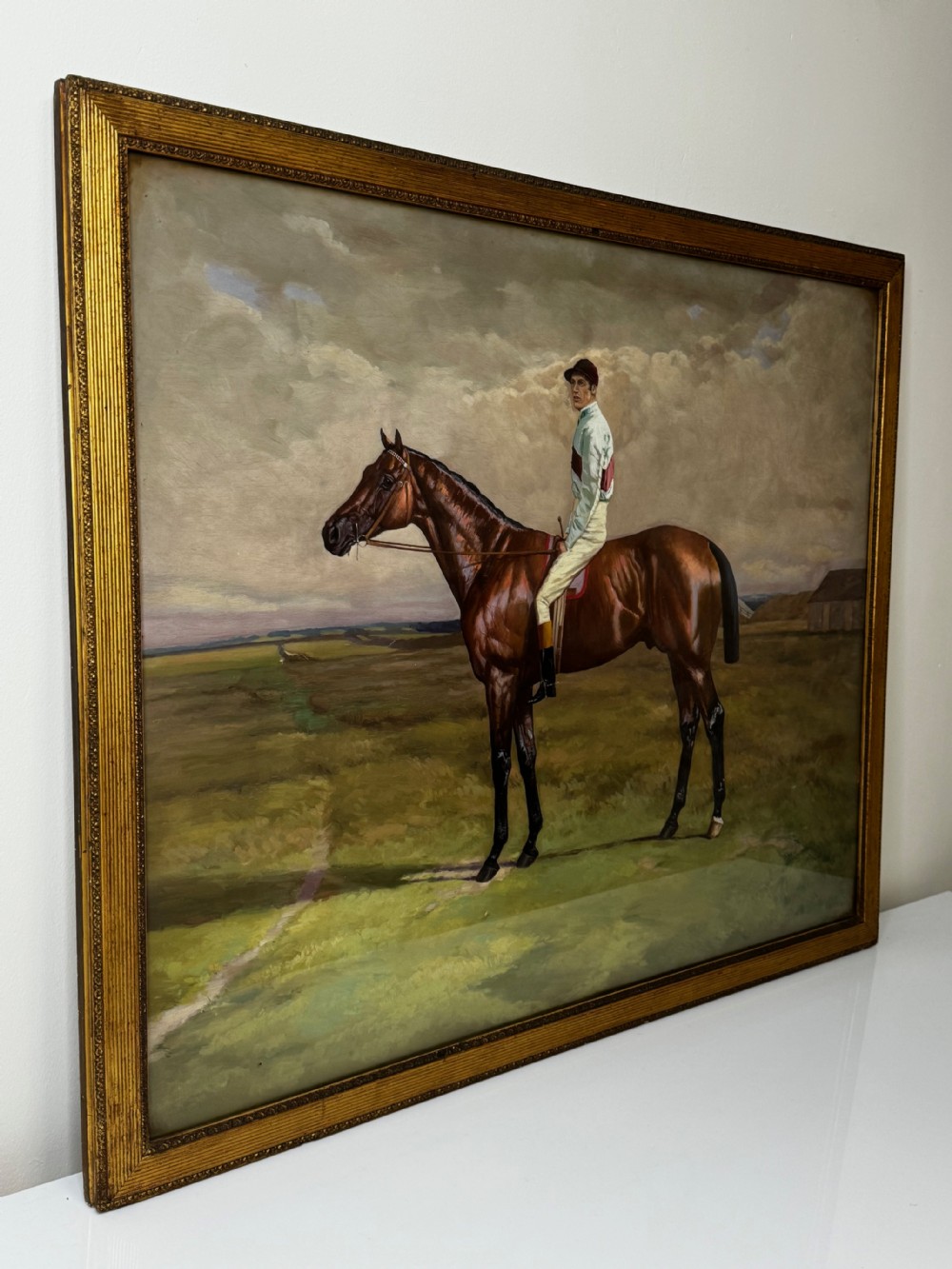 A nineteenth-century oil painting depicting a brown thoroughbred horse with a jockey in white and red silks, framed in a ribbed gold-leaf frame. View II