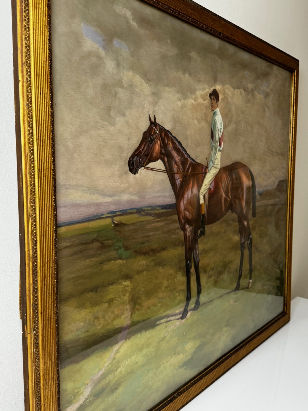 A nineteenth-century oil painting depicting a brown thoroughbred horse with a jockey in white and red silks, framed in a ribbed gold-leaf frame. View IV