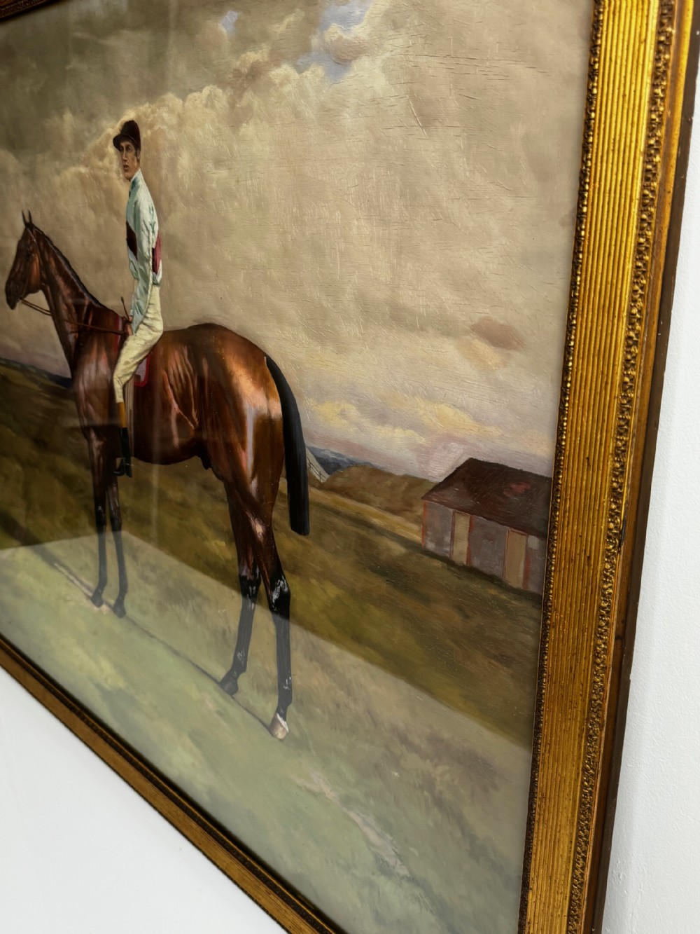 A nineteenth-century oil painting depicting a brown thoroughbred horse with a jockey in white and red silks, framed in a ribbed gold-leaf frame. View X