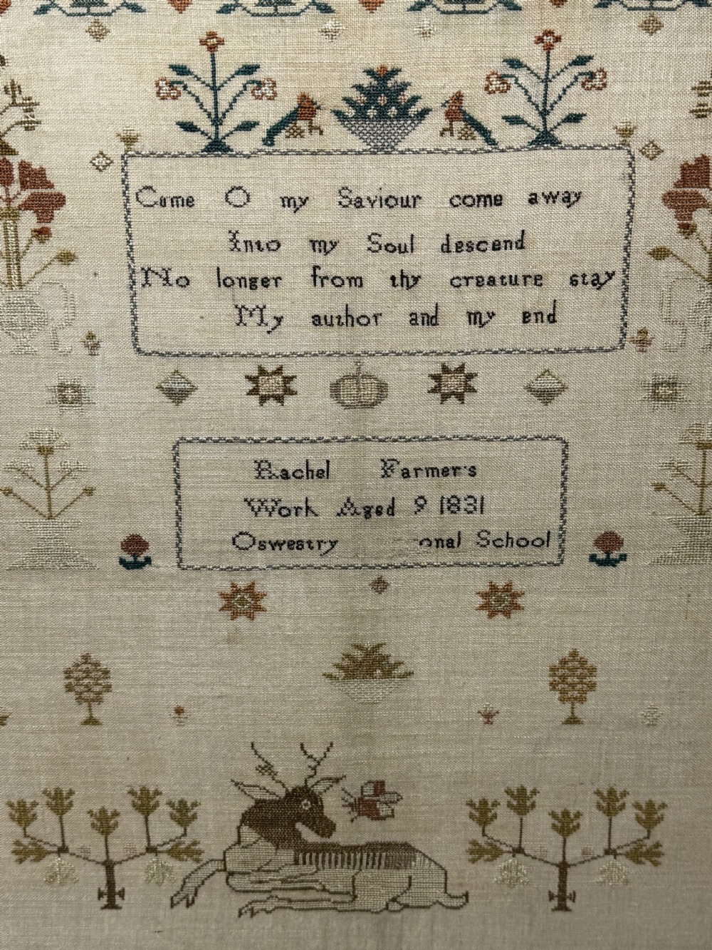 A framed 1831 needlework sampler featuring embroidered religious verse, floral borders, and a central stag motif on a linen background. View XII
