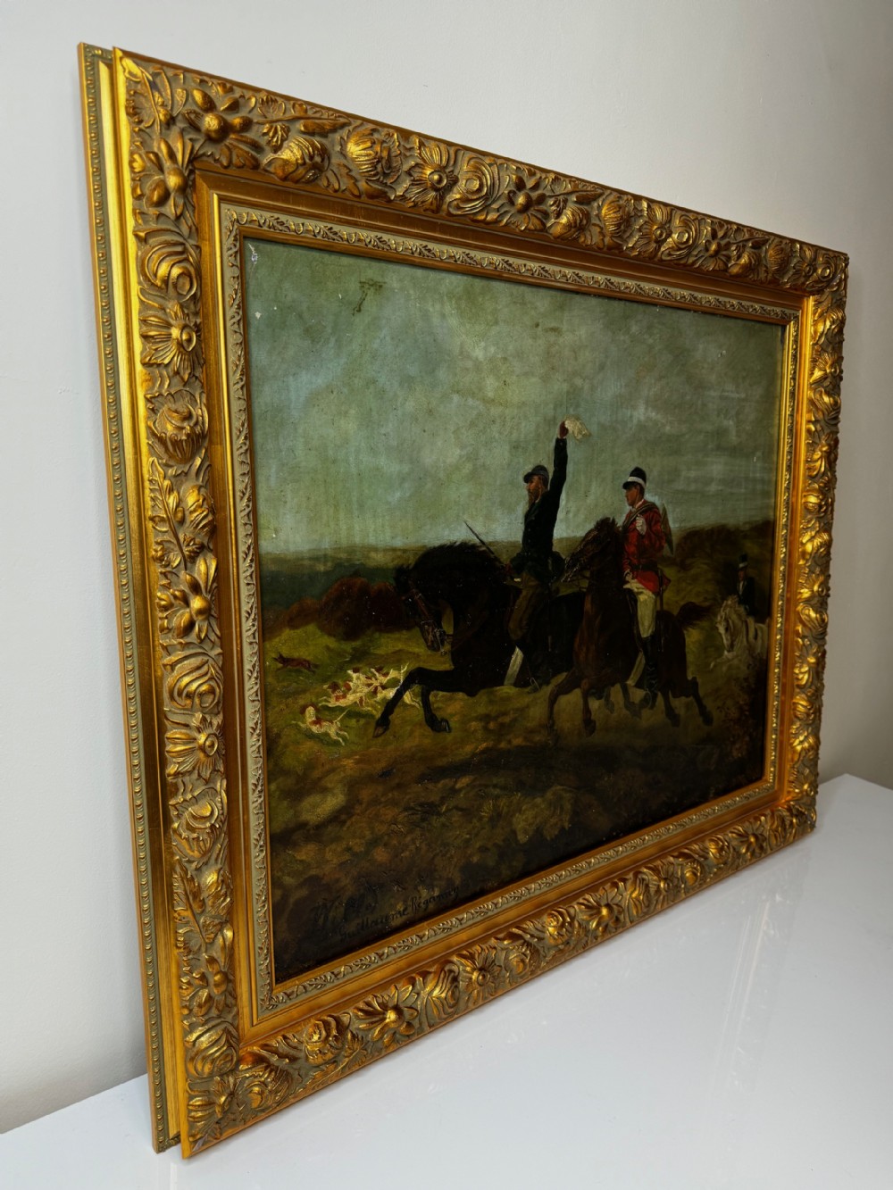 An antique oil painting depicting huntsmen riding horses alongside hounds in an open landscape, housed within an ornate carved gold gilt frame. View V
