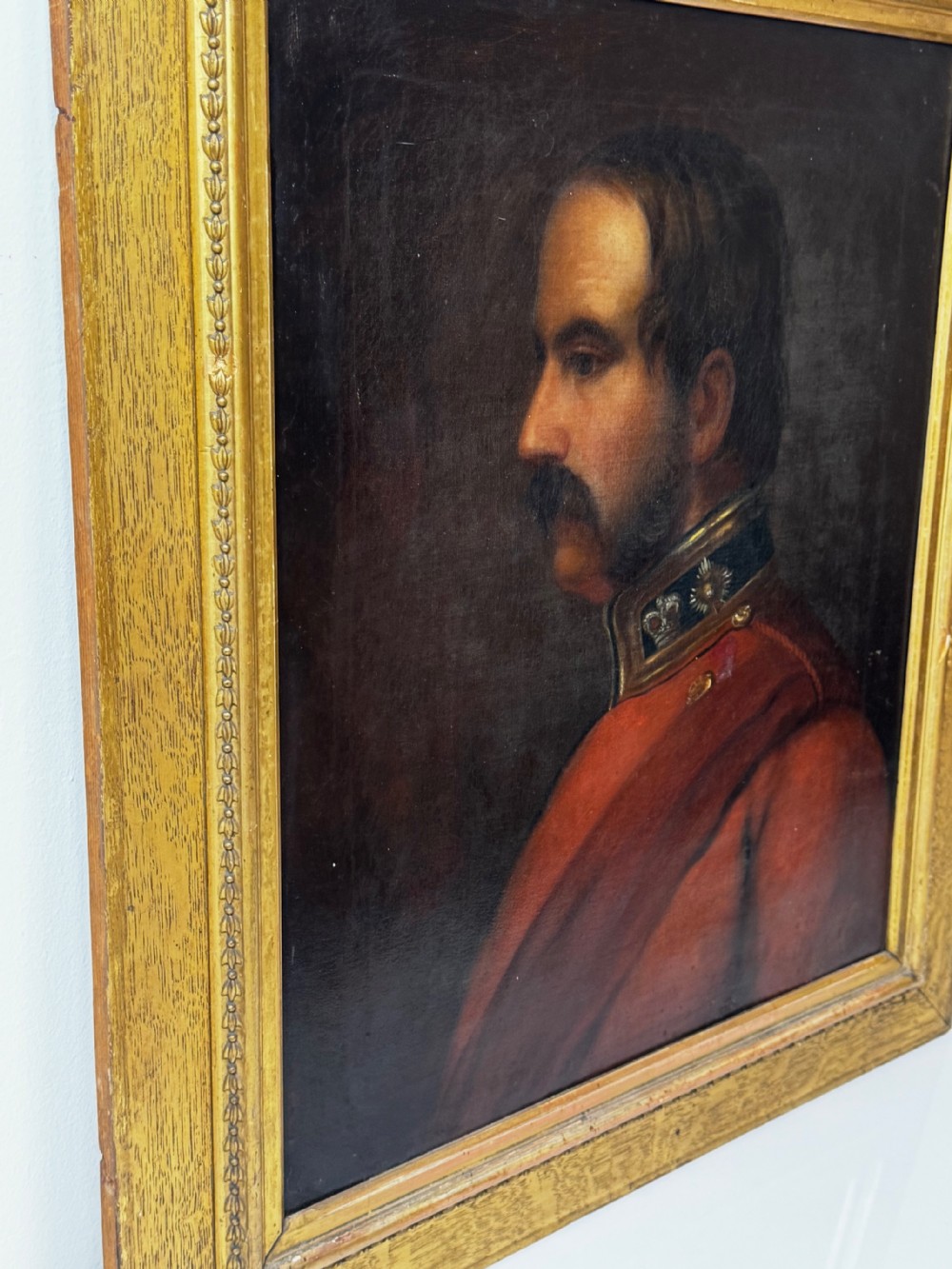 A profile view Victorian oil painting depicting a military officer with a moustache in a red uniform coat, set within a gilded wooden frame. View IX