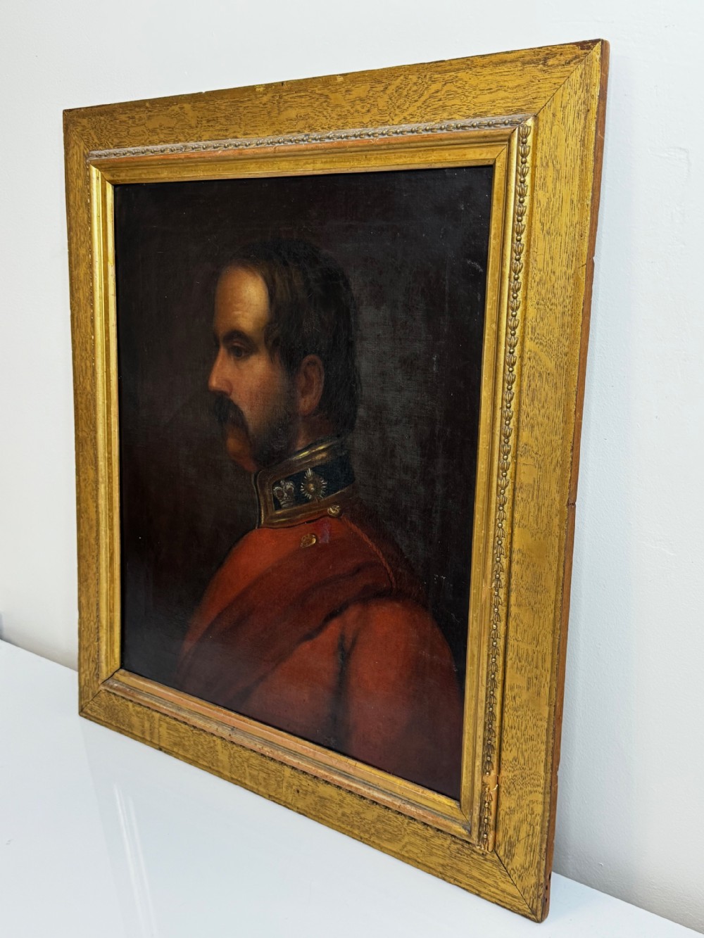 A profile view Victorian oil painting depicting a military officer with a moustache in a red uniform coat, set within a gilded wooden frame. View X