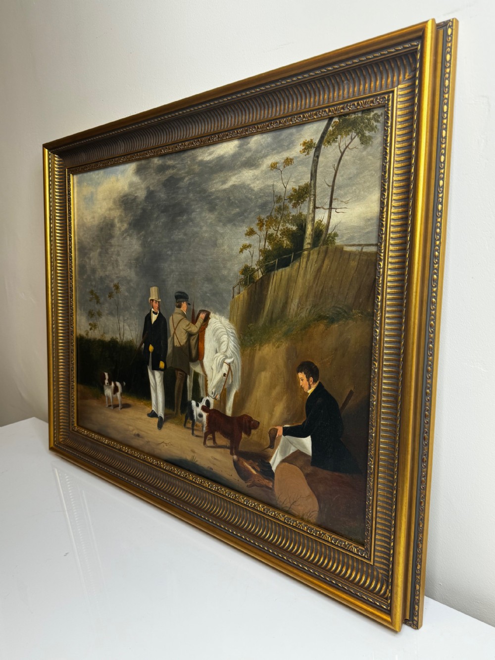 An antique oil painting in a decorative gilded frame depicting three men with hunting dogs and a white horse in a countryside setting. View X