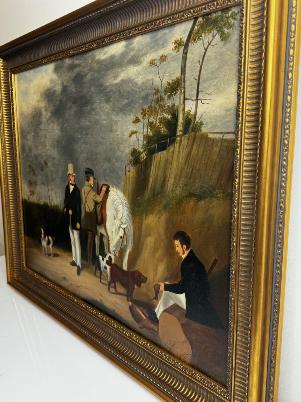 An antique oil painting in a decorative gilded frame depicting three men with hunting dogs and a white horse in a countryside setting. View XII