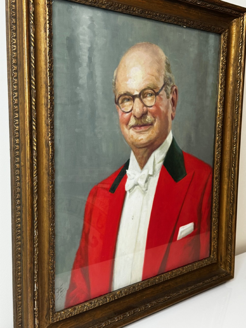 A mid-century oil painting depicting a gentleman wearing glasses, a red coat with dark lapels, and a white bow tie, housed within a gilded wooden frame. View V