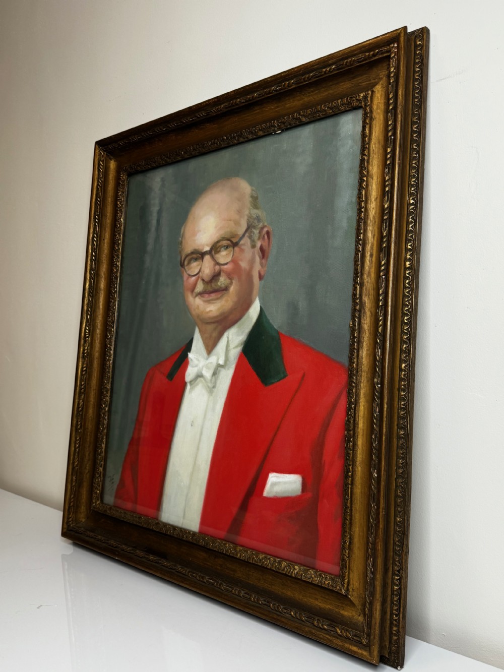 A mid-century oil painting depicting a gentleman wearing glasses, a red coat with dark lapels, and a white bow tie, housed within a gilded wooden frame. View VI