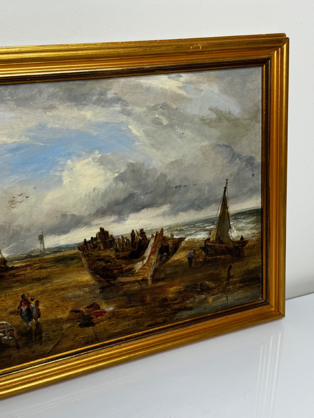 An antique oil painting depicting a grounded wooden ship wreck on a sandy shoreline with a distant lighthouse, set in a gold gilded frame. View II