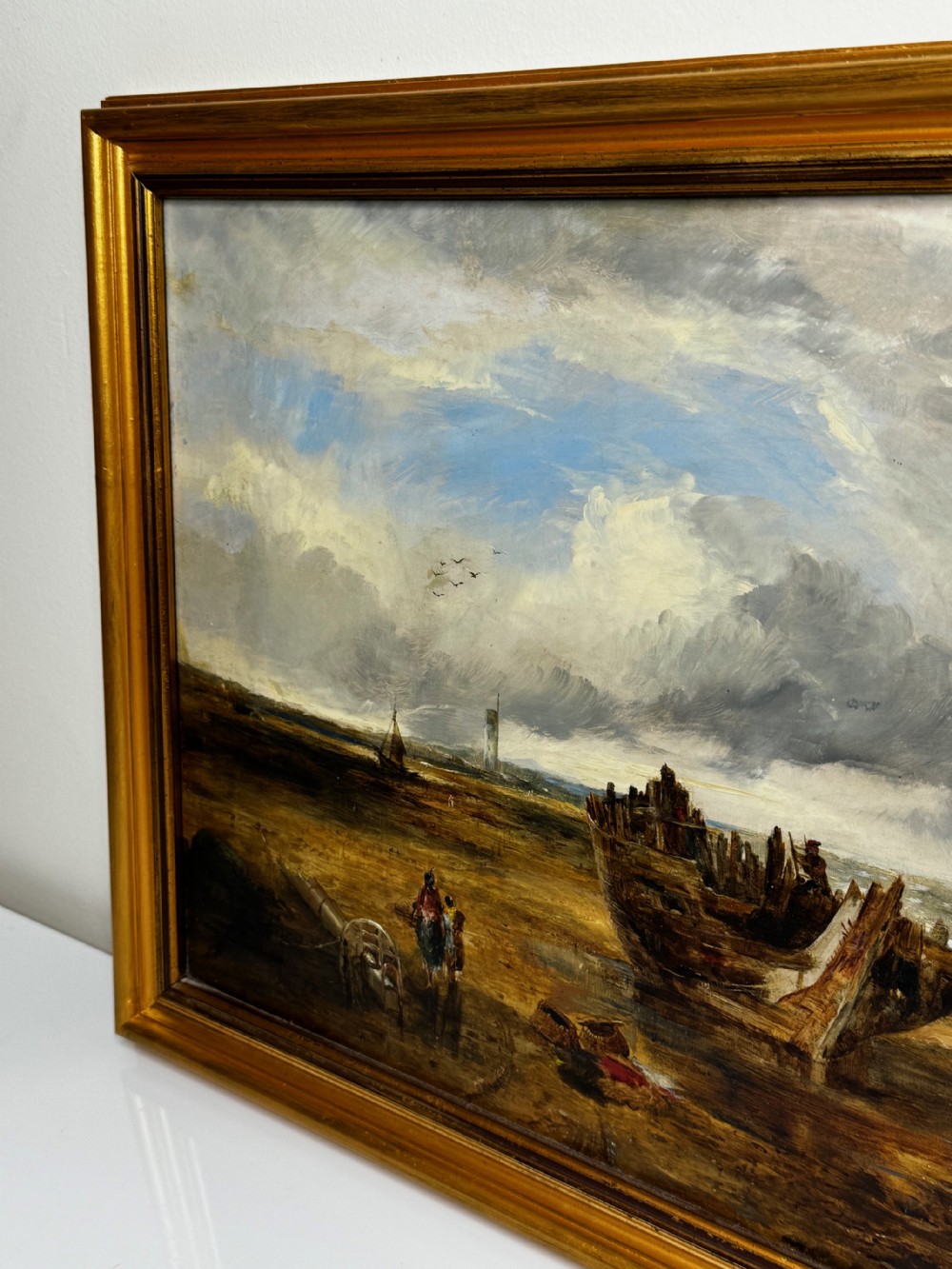 An antique oil painting depicting a grounded wooden ship wreck on a sandy shoreline with a distant lighthouse, set in a gold gilded frame. View IV