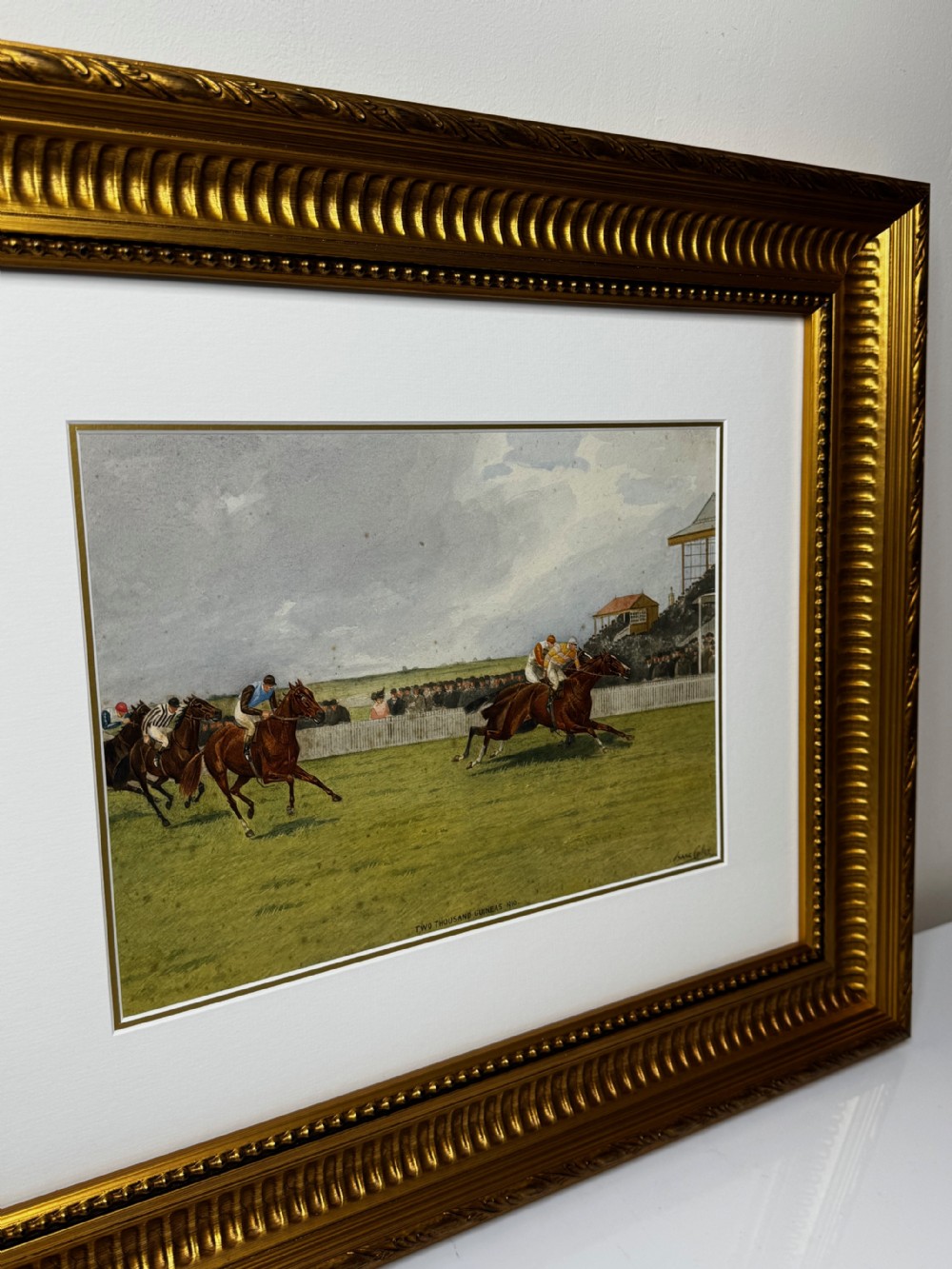 A framed British equestrian painting depicting racehorses and jockeys galloping on a green racetrack at Newmarket within a fluted gold-gilt frame. View II