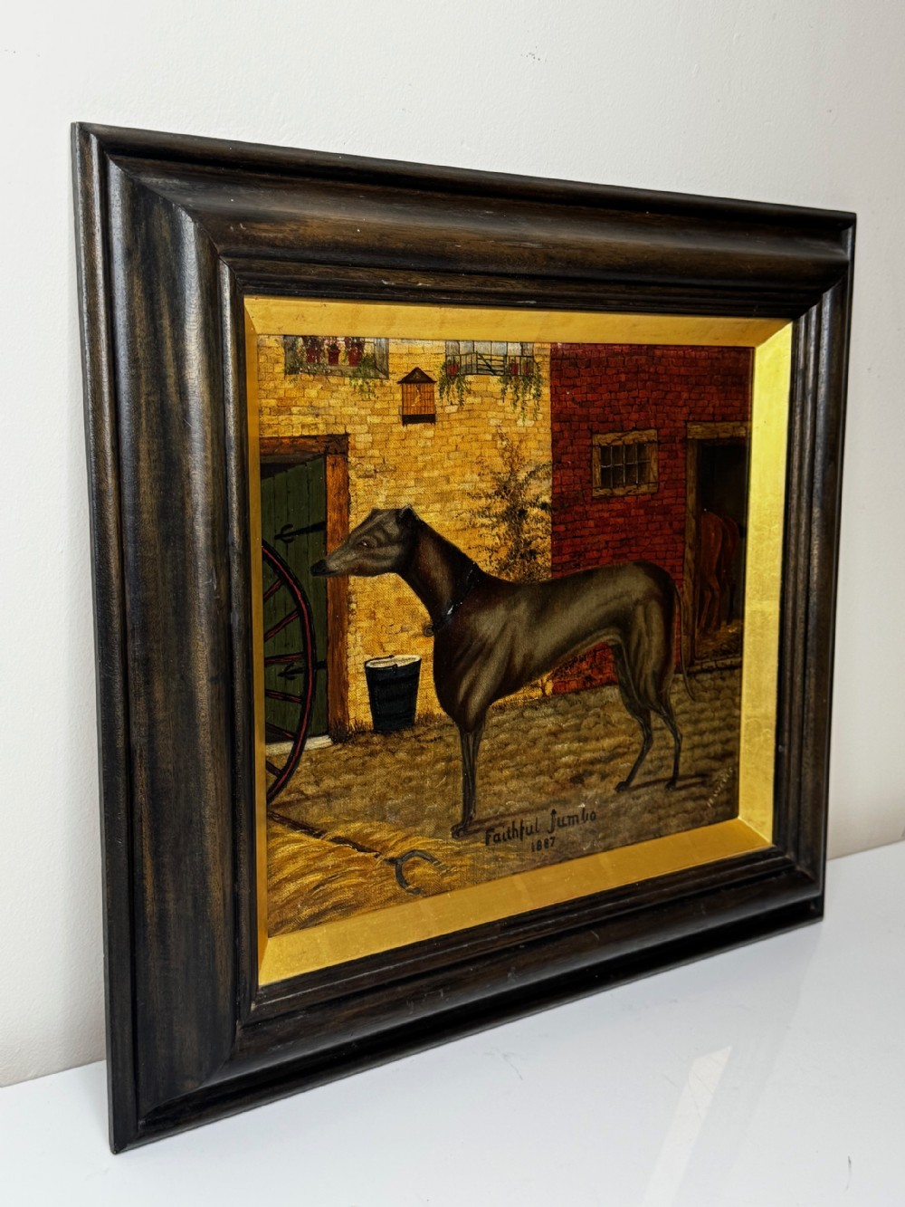 A Victorian oil painting depicting a dark brown greyhound standing in profile within a courtyard, presented in a dark molded wooden frame with a gold slip. View III