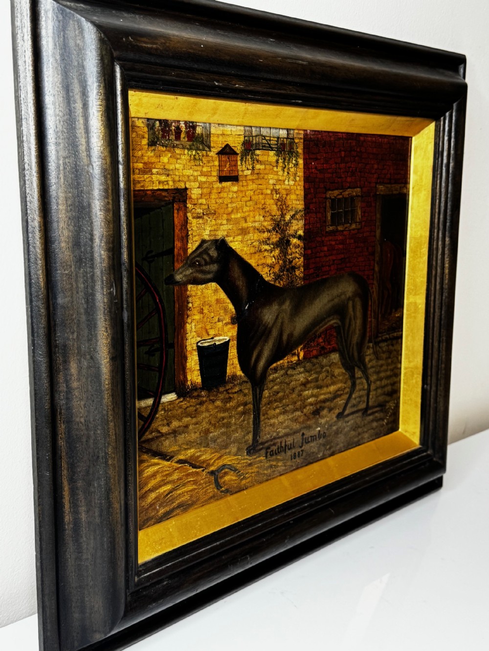 A Victorian oil painting depicting a dark brown greyhound standing in profile within a courtyard, presented in a dark molded wooden frame with a gold slip. View IV