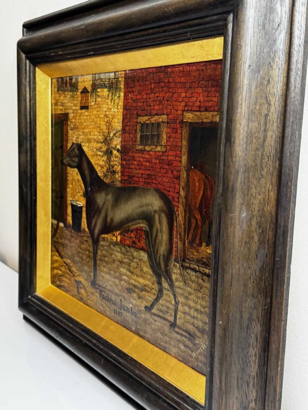 A Victorian oil painting depicting a dark brown greyhound standing in profile within a courtyard, presented in a dark molded wooden frame with a gold slip. View VII