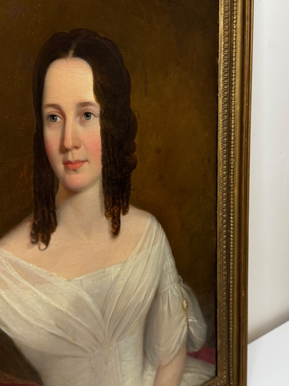 An antique Georgian oil portrait painting depicting a woman in a white off-the-shoulder gown with a red drape, framed in a beaded gold gilt frame. View II