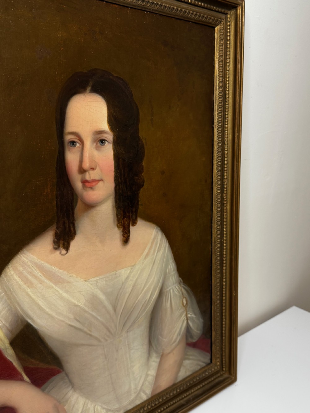 An antique Georgian oil portrait painting depicting a woman in a white off-the-shoulder gown with a red drape, framed in a beaded gold gilt frame. View III