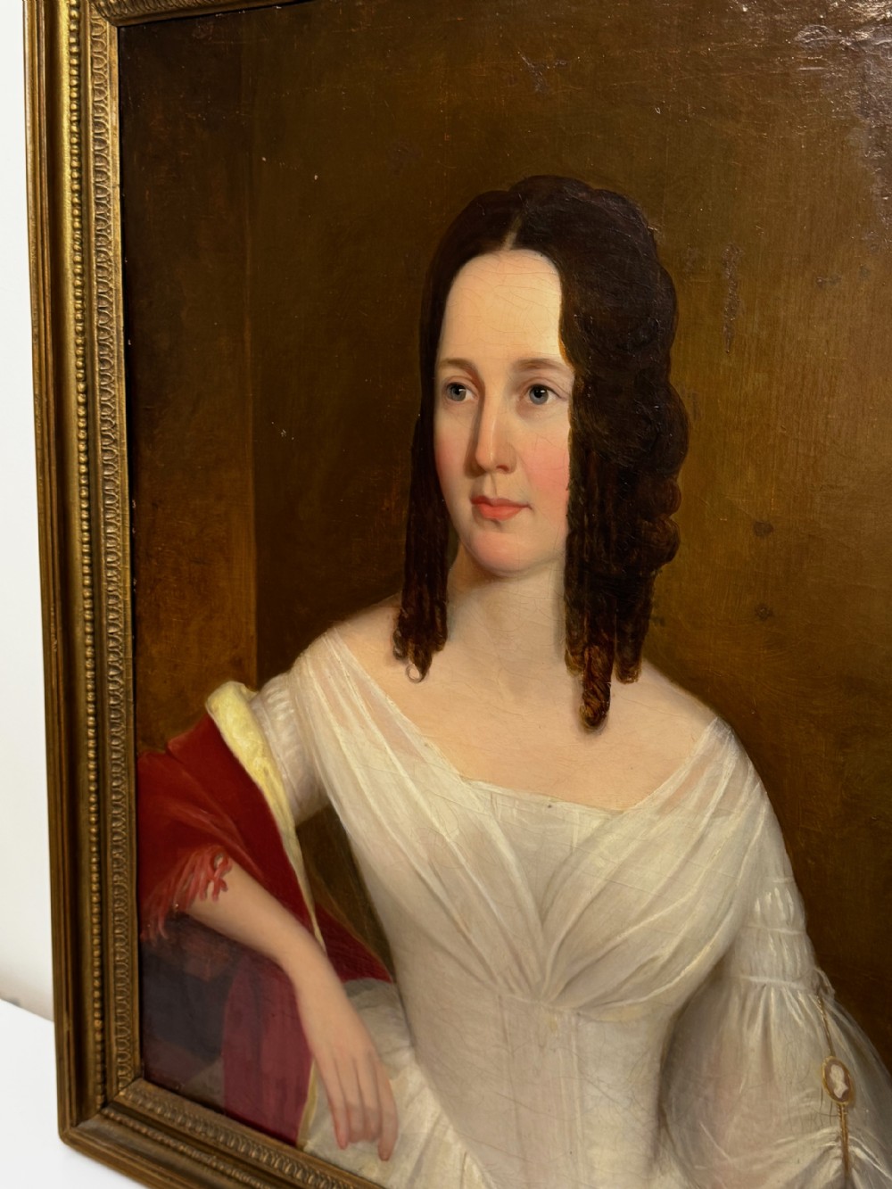 An antique Georgian oil portrait painting depicting a woman in a white off-the-shoulder gown with a red drape, framed in a beaded gold gilt frame. View IV