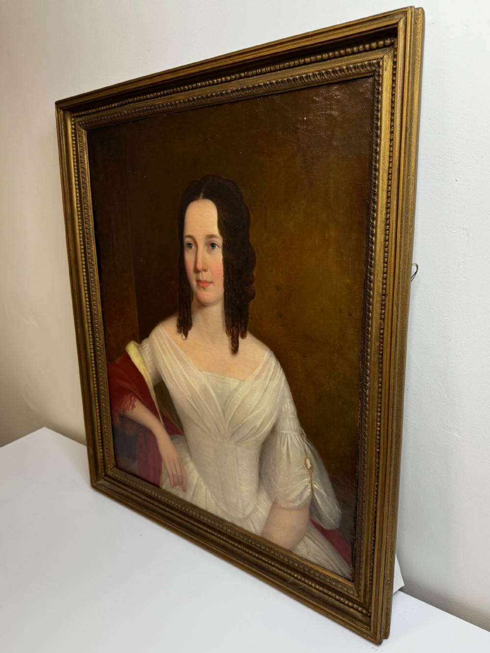 An antique Georgian oil portrait painting depicting a woman in a white off-the-shoulder gown with a red drape, framed in a beaded gold gilt frame. View V