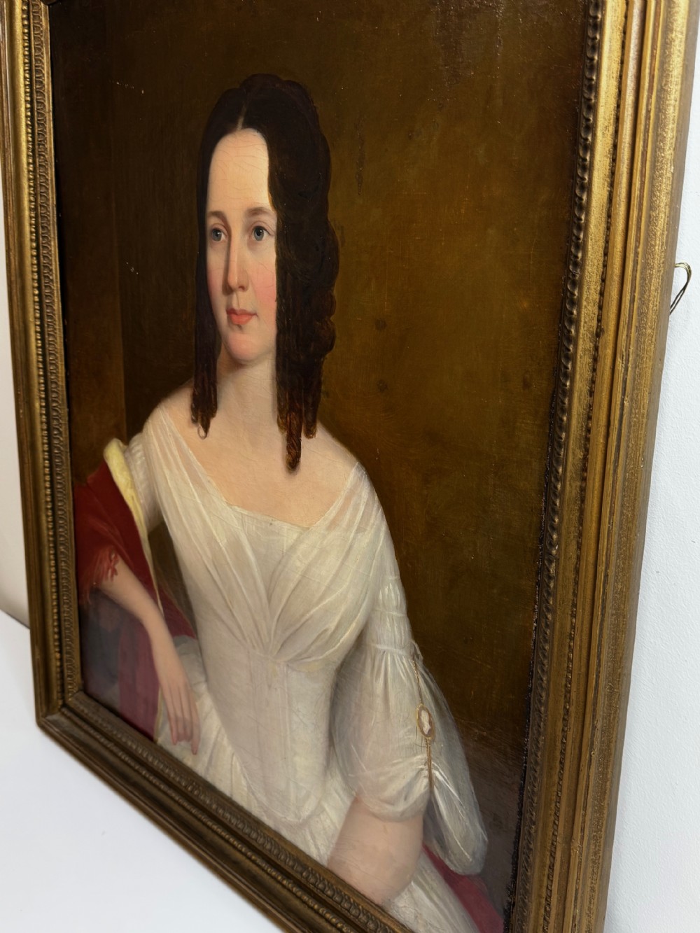 An antique Georgian oil portrait painting depicting a woman in a white off-the-shoulder gown with a red drape, framed in a beaded gold gilt frame. View VI