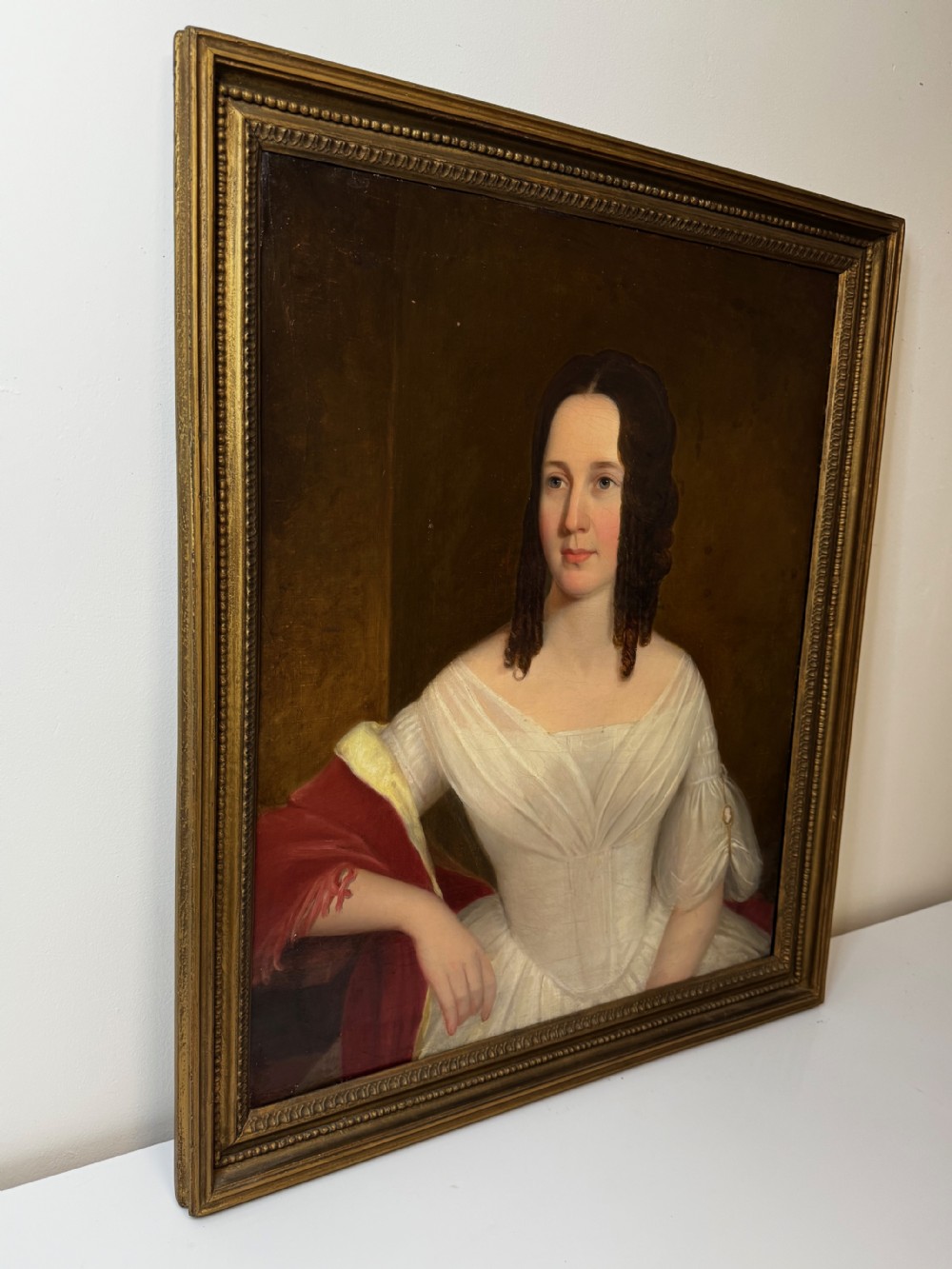 An antique Georgian oil portrait painting depicting a woman in a white off-the-shoulder gown with a red drape, framed in a beaded gold gilt frame. View X