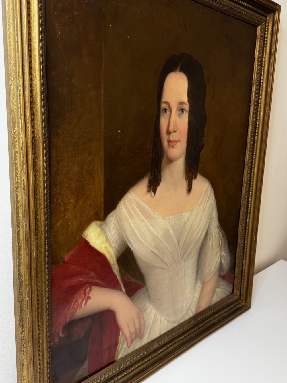 An antique Georgian oil portrait painting depicting a woman in a white off-the-shoulder gown with a red drape, framed in a beaded gold gilt frame. View XI