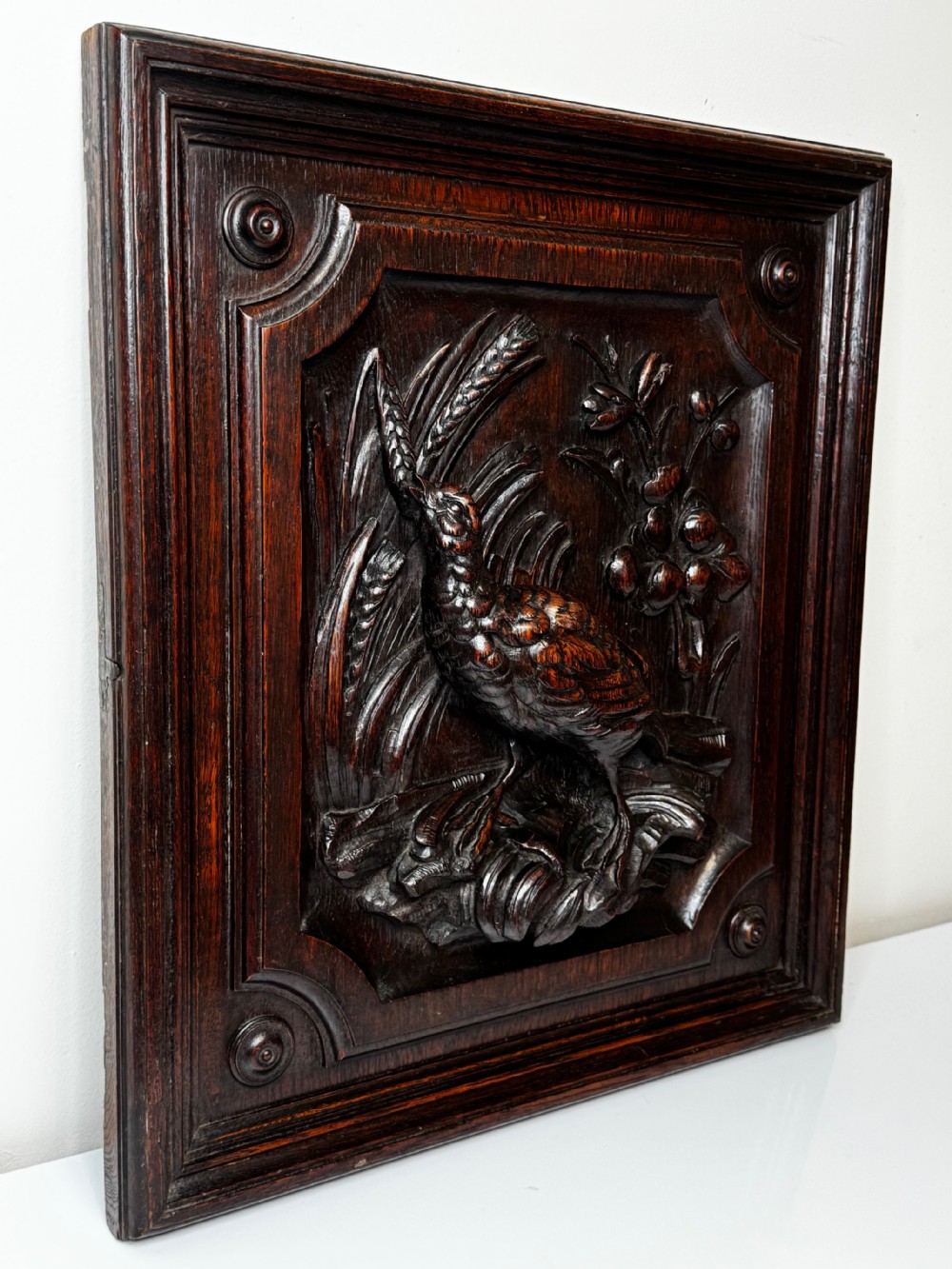A dark oak relief carving depicting a game bird standing among reeds and oak leaves with acorns inside a moulded frame. View II