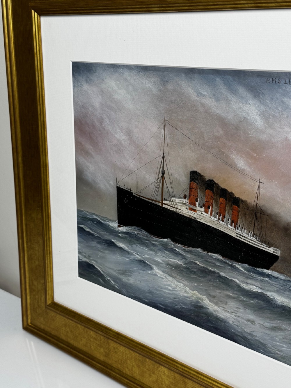 A framed marine painting depicting the RMS Lusitania ocean liner navigating choppy seas under a cloudy sky, enclosed within a textured gilt frame. View III