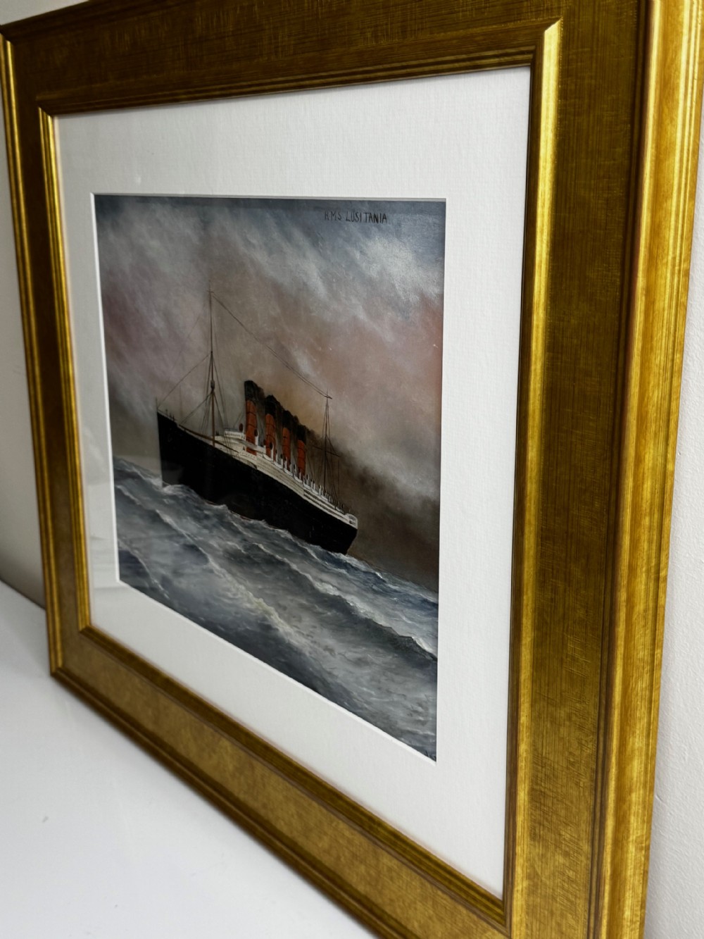 A framed marine painting depicting the RMS Lusitania ocean liner navigating choppy seas under a cloudy sky, enclosed within a textured gilt frame. View IV