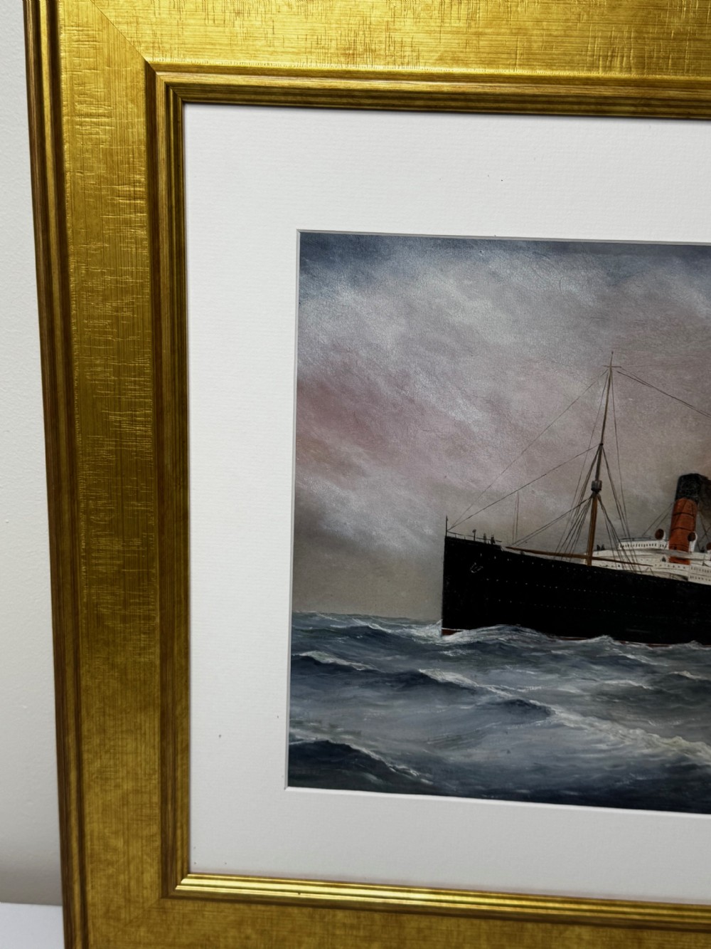 A framed marine painting depicting the RMS Lusitania ocean liner navigating choppy seas under a cloudy sky, enclosed within a textured gilt frame. View VI