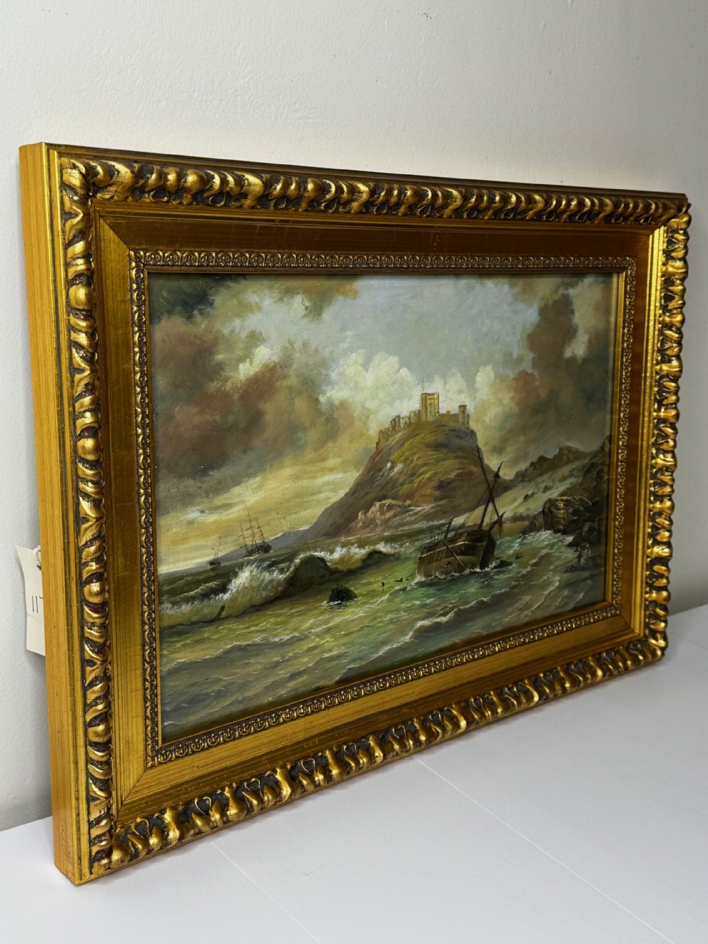 A Victorian oil painting depicting a coastal shipwreck near Bamburgh Castle under a cloudy sky, enclosed in a heavily gilded frame. View VI