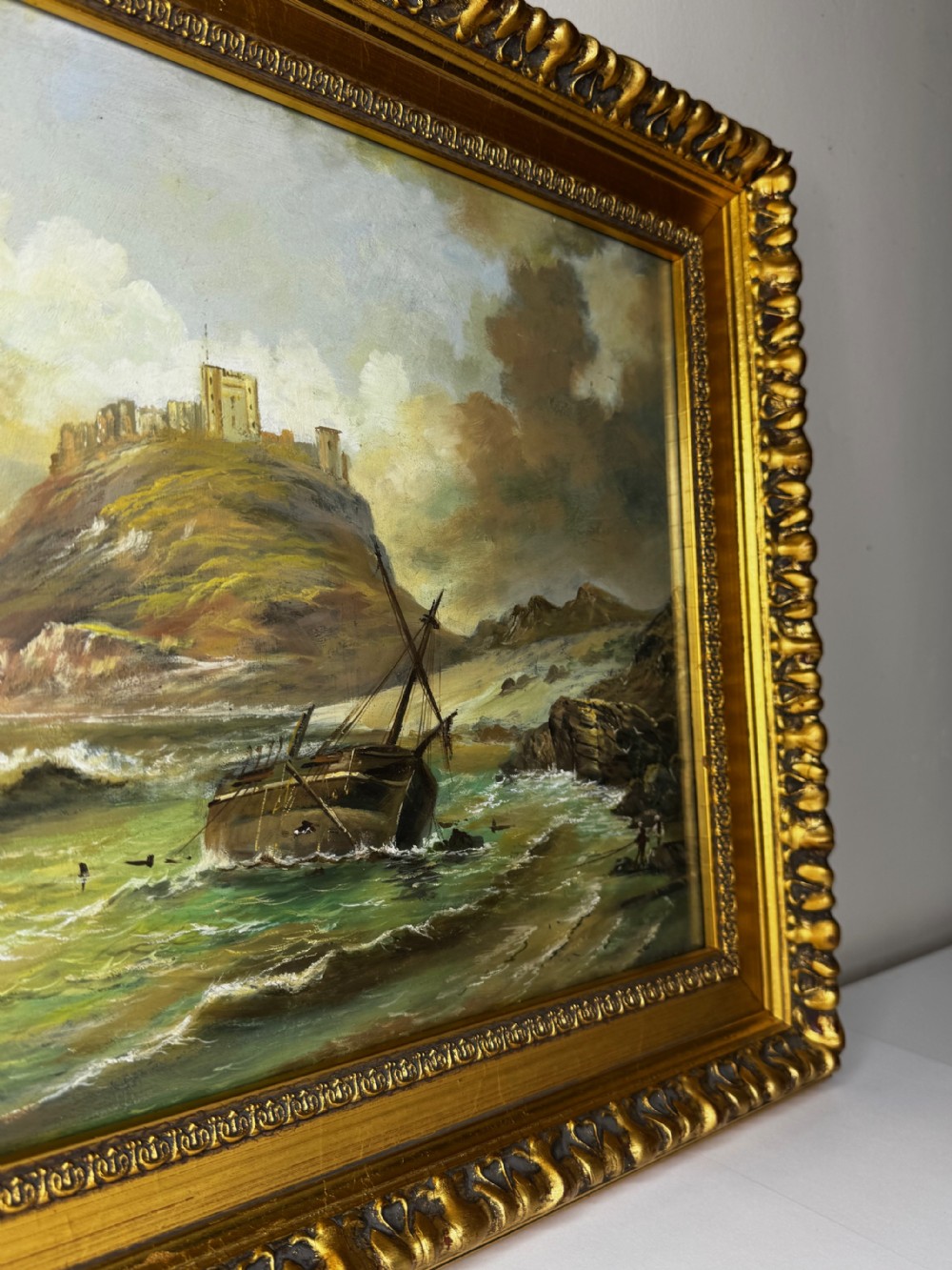 A Victorian oil painting depicting a coastal shipwreck near Bamburgh Castle under a cloudy sky, enclosed in a heavily gilded frame. View XIV