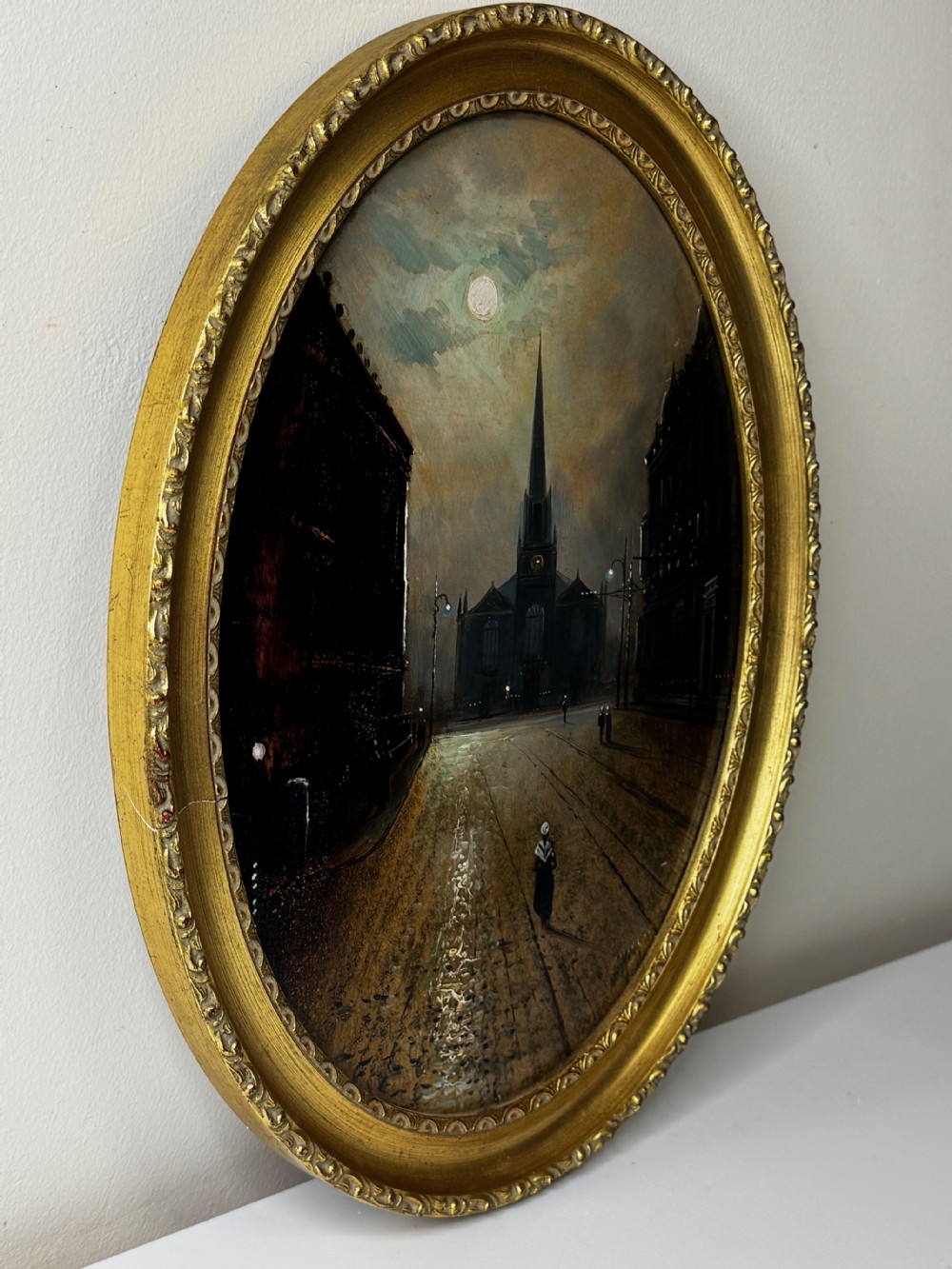 An oval Victorian oil painting depicting a wet night street lit by moonlight leading towards a church spire, framed in a carved gilt frame. View VI