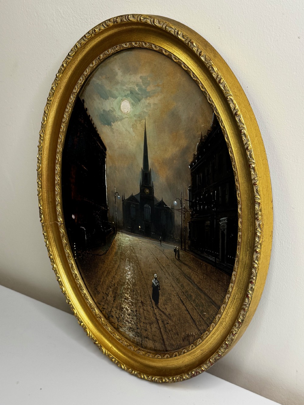 An oval Victorian oil painting depicting a wet night street lit by moonlight leading towards a church spire, framed in a carved gilt frame. View VIII
