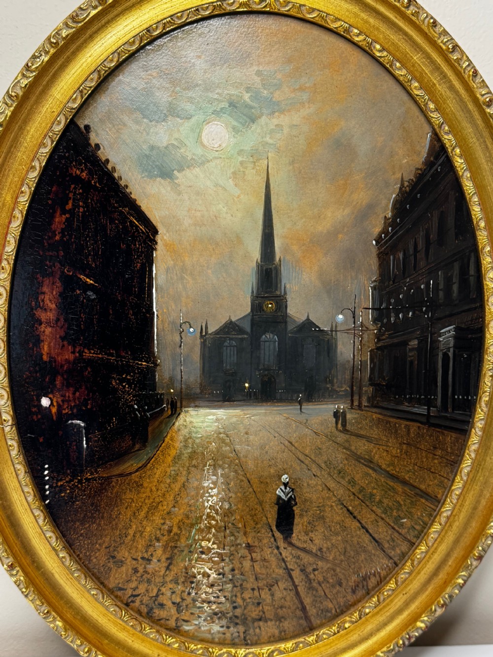 An oval Victorian oil painting depicting a wet night street lit by moonlight leading towards a church spire, framed in a carved gilt frame. View X