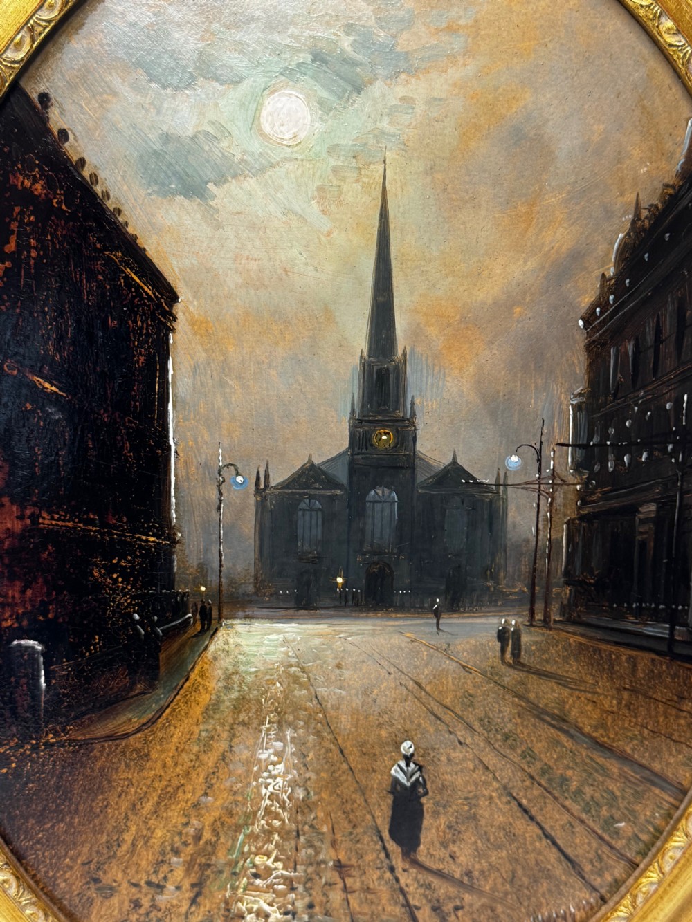 An oval Victorian oil painting depicting a wet night street lit by moonlight leading towards a church spire, framed in a carved gilt frame. View XI